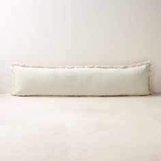 a white rectangular pillow with fringe trim on the bottom and sides, sitting against a wall