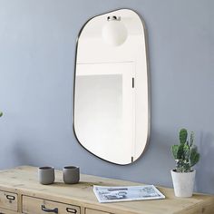 a mirror on the wall above a dresser with two mugs and a potted plant