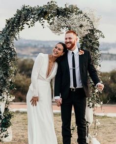 a man and woman are standing under an arch with greenery on the sides, smiling