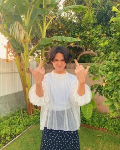 a woman standing in front of a tree making the peace sign with her hands while wearing a white top