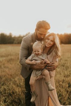 a man and woman holding a baby in a field at sunset with the sun behind them