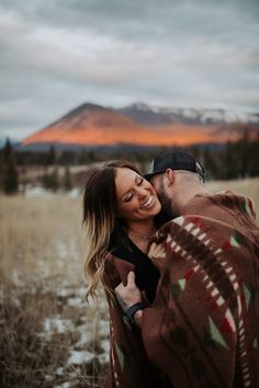 a man and woman are hugging in the field with mountains in the background at sunset