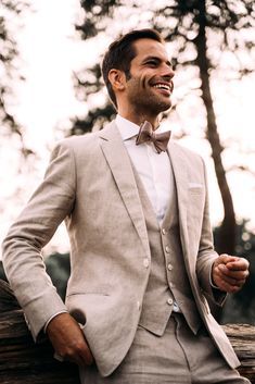 a man in a suit and bow tie sitting on a tree trunk smiling at the camera