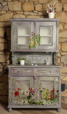 an old dresser painted with flowers and plants on the front, next to a stone wall