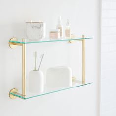 two glass shelves with toothbrushes, soap and lotion bottles on them against a white wall