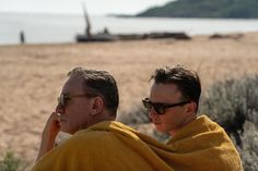 two men wrapped in yellow towels sitting on the beach looking out at the water and sand