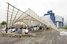 several people sitting on wooden benches in front of a large structure made out of wood planks