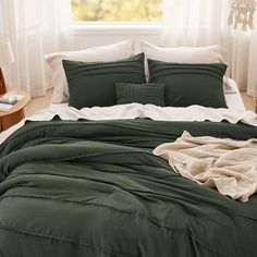 a bed with green comforter and pillows in a room next to a window that has white curtains