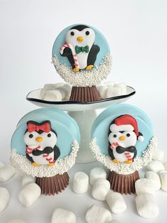 there are three cupcakes decorated with penguins on top of the cake stand and white marshmallows scattered around them