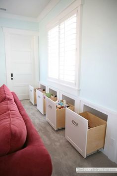 a red couch sitting in front of a window next to two bins filled with toys