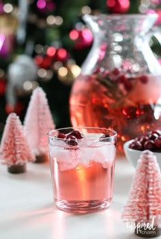 a glass filled with cranberry shrub next to a pitcher of liquid and small trees