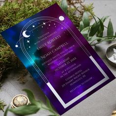 a purple and blue wedding card sitting on top of a table next to greenery