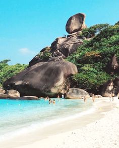 people are swimming in the clear blue water near large rocks and boulders on a white sandy beach