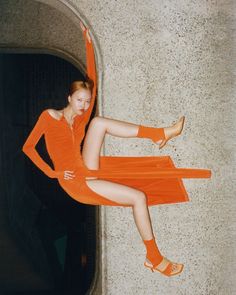 a woman in an orange dress is leaning against a wall with her legs spread out