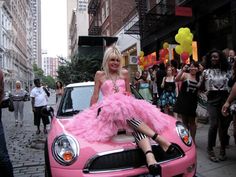 a woman in a pink dress sitting on top of a pink car with people walking around