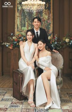 two women and a man sitting on a chair in front of a chandelier