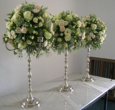 three silver vases filled with flowers on top of a white marble table next to a bookshelf