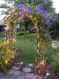 a garden with purple flowers and lights on the arbor in front of it is surrounded by greenery