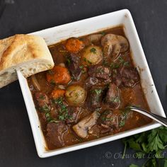 a bowl of beef stew with bread and parsley garnish on the side