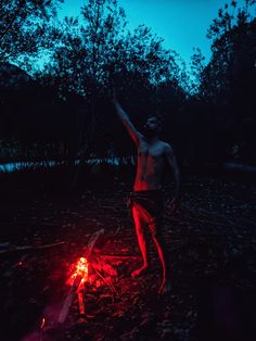 a man standing next to a campfire with his arms up in the air at night