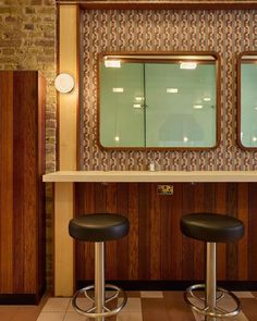 two stools in front of a counter with mirrors on the wall and tiled floor