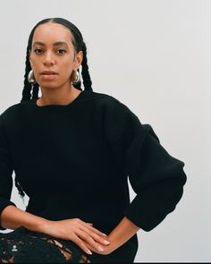 a woman with braids standing in front of a white wall wearing a black sweater