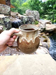 a person holding a vase in front of a river with rocks and trees around it