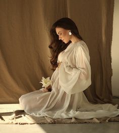 a pregnant woman sitting on the floor with flowers in her hand and wearing a white dress