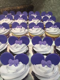 cupcakes with white frosting and purple bows are arranged in rows on a table