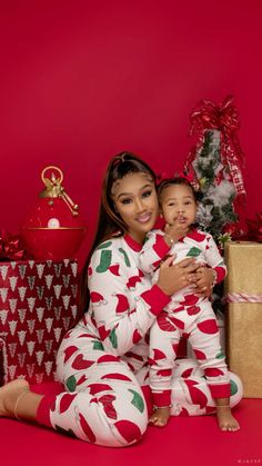 a woman in pajamas holding a baby next to christmas presents on a red background,