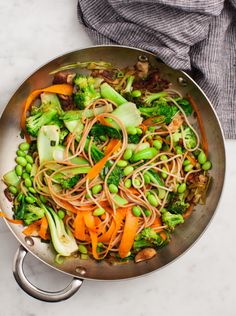 a wok filled with vegetables and noodles on top of a white counter next to a gray towel