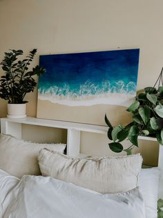 a painting on the wall above a bed with white sheets and pillows, next to a potted plant