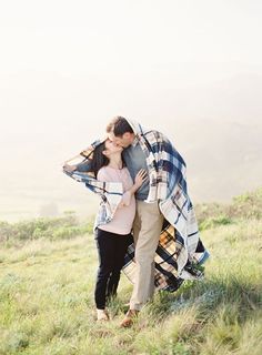 a man and woman kissing under a blanket on top of a grass covered hill with mountains in the background