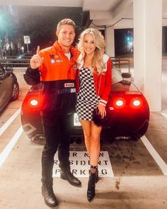 a man and woman standing next to a car in a parking lot with their thumbs up