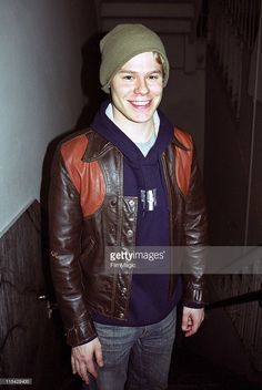 a young man wearing a brown leather jacket and a green beanie smiles at the camera