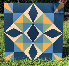 a hand is holding up a blue and yellow quilt on the grass in front of a chain link fence