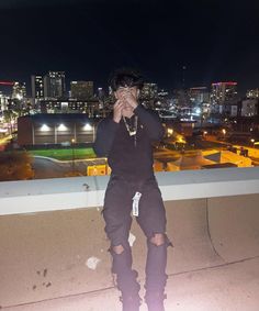 a man sitting on top of a building talking on a cell phone with city lights in the background