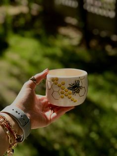 a woman holding a cup with bees on it in front of some trees and bushes