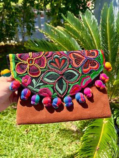 a hand holding a colorful embroidered clutch bag in front of a palm tree and green grass