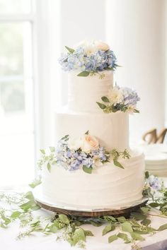 a three tiered cake with blue and white flowers on the top is surrounded by greenery