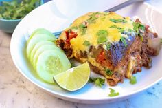 a white plate topped with an enchilada covered in cheese next to sliced cucumbers