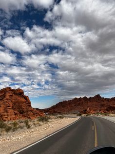 a car driving down the road in front of some red rocks and mountains under a cloudy sky