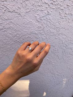 a person's hand with a diamond ring on their left index finger, against a white painted wall