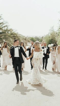 a bride and groom walking with their bridal party in the background, all dressed in black tuxedos