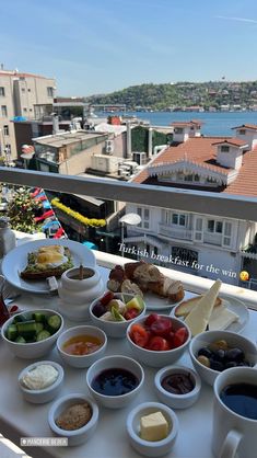 a table topped with bowls of food next to a balcony overlooking the water and buildings