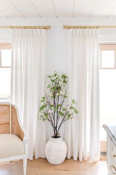 a white vase with flowers in it sitting next to a chair and window covered by curtains