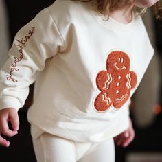 Close-up of a child wearing a beige sweater with a large gingerbread man patch in orange-brown on the front. The phrase "gingerbread kisses" is embroidered on the sleeve in cursive, enhancing the holiday feel. Hand Drawn Design