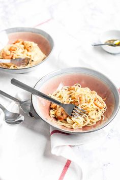 two bowls filled with pasta and shrimp on top of a tablecloth covered table next to utensils