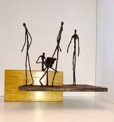 three metal figures are standing on top of a piece of wood in front of a white wall