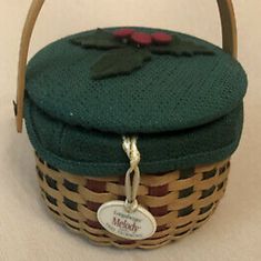 a green and brown basket with a red flower on it's top, sitting on a white surface